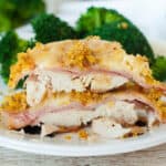 Sliced chicken cordon bleu on a white plate, showcasing layers of chicken, ham, and cheese, topped with a golden breadcrumb crust. Fresh broccoli is visible in the background, complementing the dish.