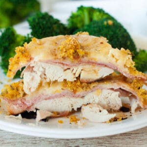 Sliced chicken cordon bleu on a white plate, showcasing layers of chicken, ham, and cheese, topped with a golden breadcrumb crust. Fresh broccoli is visible in the background, complementing the dish.