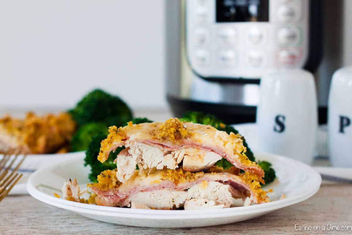 A plate with sliced chicken cordon bleu showcases layers of breaded chicken, ham, and cheese. In the background, an instant pot hints at the quick preparation of this delightful recipe alongside a piece of broccoli, a fork, and salt and pepper shakers.
