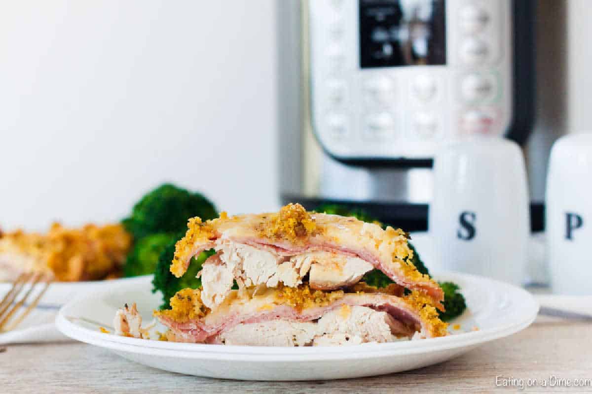 A white plate displays a sliced chicken cordon bleu with layers of ham, cheese, and a golden breadcrumb crust. An Instant Pot sits nearby, suggesting this delightful dish was crafted using an Instant Pot recipe. Broccoli and salt and pepper shakers complete the scene.
