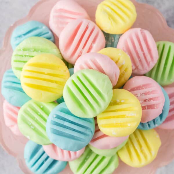 A plate filled with assorted pastel-colored cream cheese mints, featuring pink, yellow, green, and blue hues. Each piece is patterned with parallel grooves on top, creating a visually appealing texture.