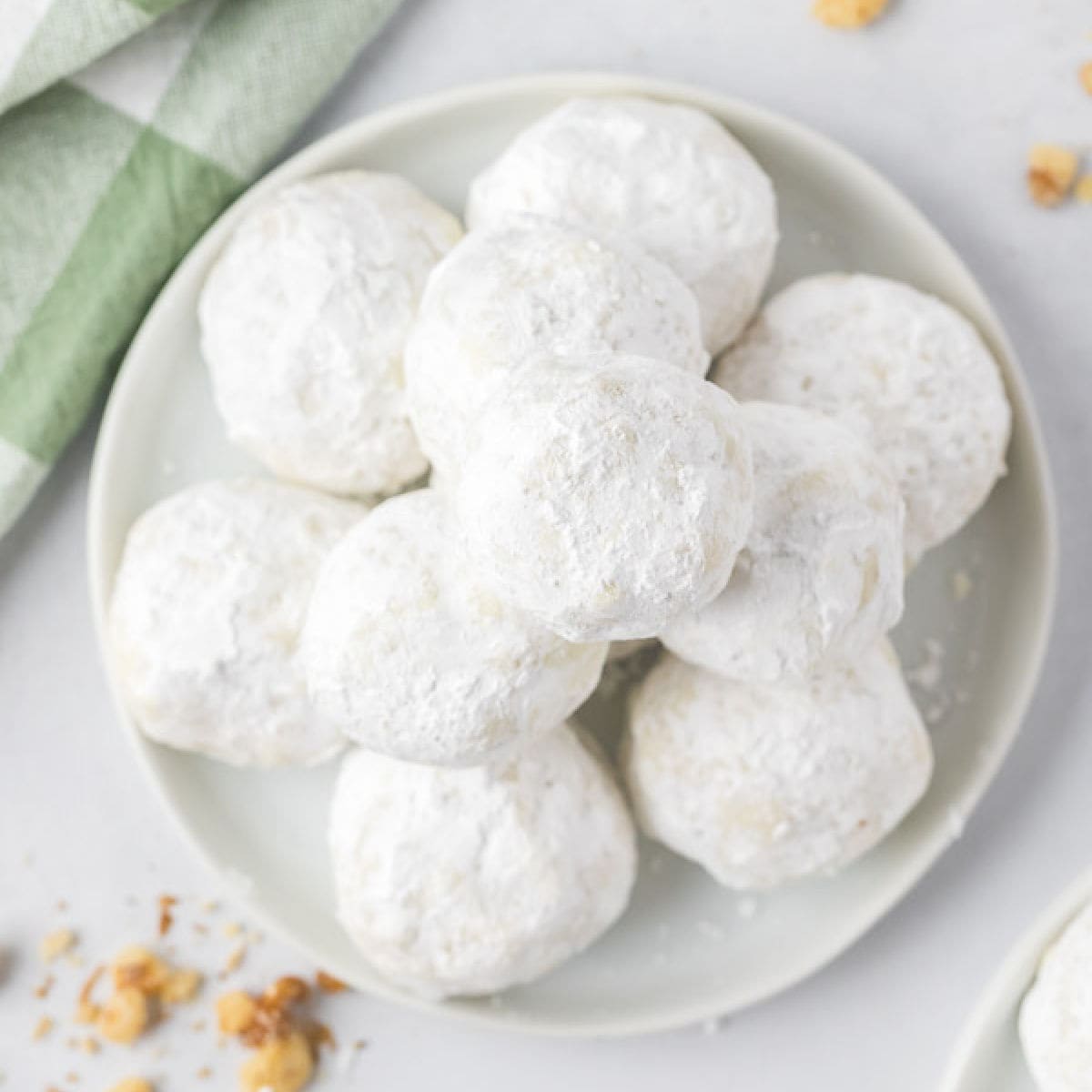 A plate of round Russian Tea Cakes, dusted with powdered sugar, is stacked invitingly on a light-colored dish. A green and white cloth peeks from the background, while crumbs are scattered nearby, hinting at their deliciousness.