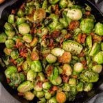A close-up of Bacon Brussel Sprouts mixed with crispy bacon pieces in a black skillet. The vegetables are charred and golden brown, with visible herbs and seasonings for extra flavor.