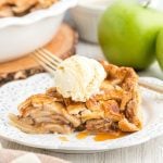A slice of Salted Caramel Apple Pie with a golden crust is topped with vanilla ice cream and caramel drizzle, served on a decorative white plate. Green apples and a pie dish are visible in the background.