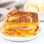 A Grilled Ham and Cheese Sandwich with melted cheddar oozing out is stacked on a white plate. A bowl of potato chips is blurred in the background.