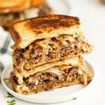 A close-up of two stacked Patty Melt sandwiches, each filled with ground beef, caramelized onions, and melted cheese between toasted slices of bread, served on a white plate.