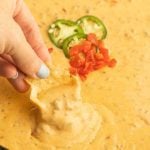 A hand dips a tortilla chip into creamy Rotel Dip, garnished with sliced jalapeños and diced tomatoes.