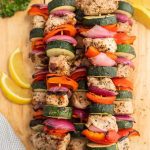 Colorful grilled pork kabobs with chunks of seasoned chicken, zucchini, red bell pepper, and red onion on skewers, arranged on a wooden board with lemon wedges on the side.