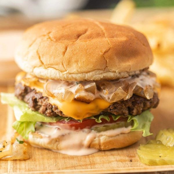 A cheeseburger with lettuce, tomato, grilled onions, melted cheese, and sauce on a bun—served In-N-Out Burger Animal Style—with pickle slices and fries in the background on a wooden surface.