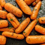 Close-up of roasted baby carrots sprinkled with chopped herbs and seasoning, arranged on a dark surface. The carrots are glistening with a slightly caramelized texture, perfect for any Ranch Carrots Recipe.