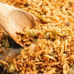 A wooden spoon scoops a serving of Skillet Green Bean Casserole topped with crispy fried onions and green beans. The dish appears golden brown and crunchy.
