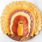A festive Thanksgiving Turkey Charcuterie Board arranged to resemble a turkey, with layers of crackers, cheese, and meats for feathers, a body made of cheese and pepperoni, and two chocolate chips for eyes.