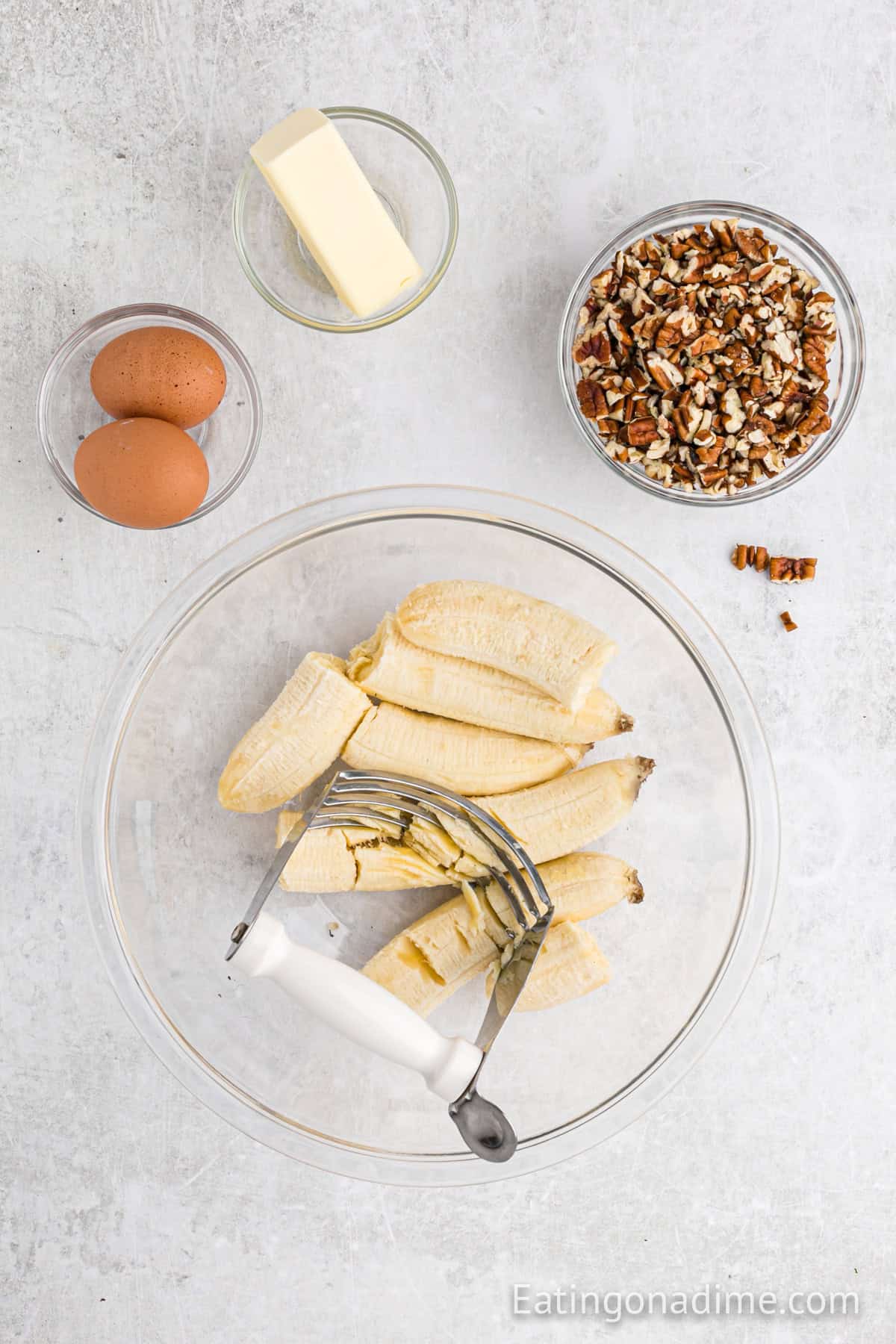 Bananas in a bowl ready to be smashed with small bowls of eggs, butter, and pecans.