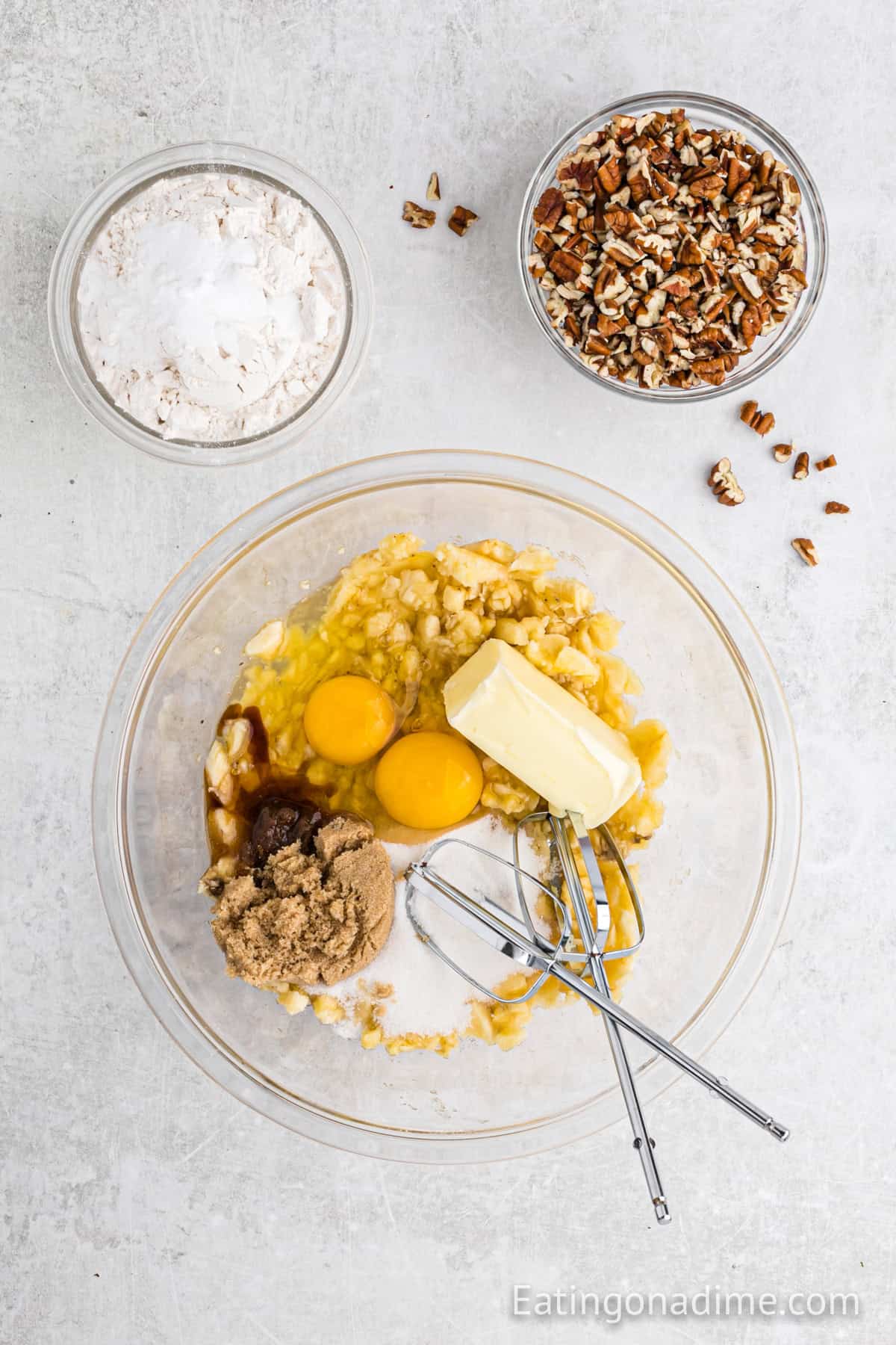 Mashed bananas, eggs, butter, brown sugar, and vanilla in a bowl. With a small bowl of flour and pecans on the side.