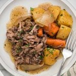 A plate of Slow Cooker Red Wine Pot Roast with shredded beef, large chunks of potatoes, carrots, onions, and rich gravy, garnished with chopped parsley.