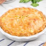 An Easy Turkey Pot Pie with a golden-brown, flaky crust sits in a white ceramic dish on a marble surface, garnished with parsley leaves and a striped cloth nearby.