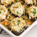 A close-up of Olive Garden Stuffed Mushrooms in a white baking dish, topped with melted cheese, breadcrumbs, and chopped parsley, served over a golden sauce.