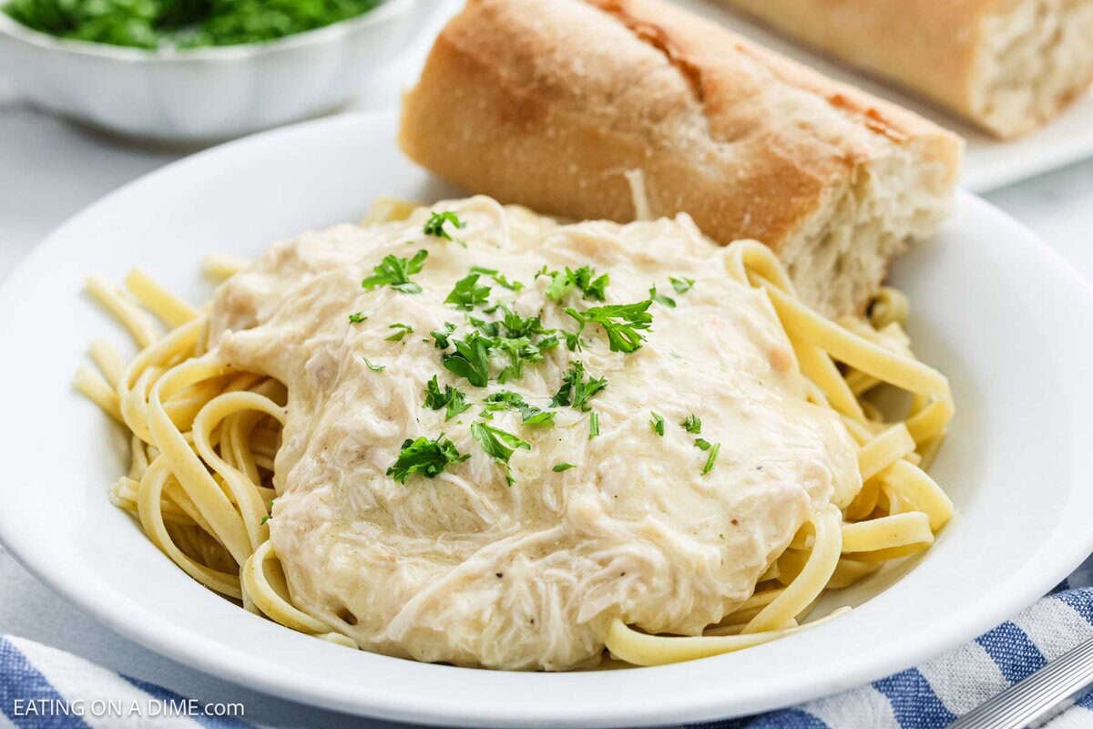 A white plate of fettuccine pasta topped with creamy Crock Pot Chicken Alfredo sauce and shredded chicken, garnished with chopped parsley. A piece of bread sits on the side, with a bowl of chopped herbs in the background.