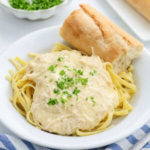 A bowl of fettuccine pasta topped with creamy Crock Pot Chicken Alfredo sauce and garnished with chopped parsley, served with a piece of crusty bread on the side.