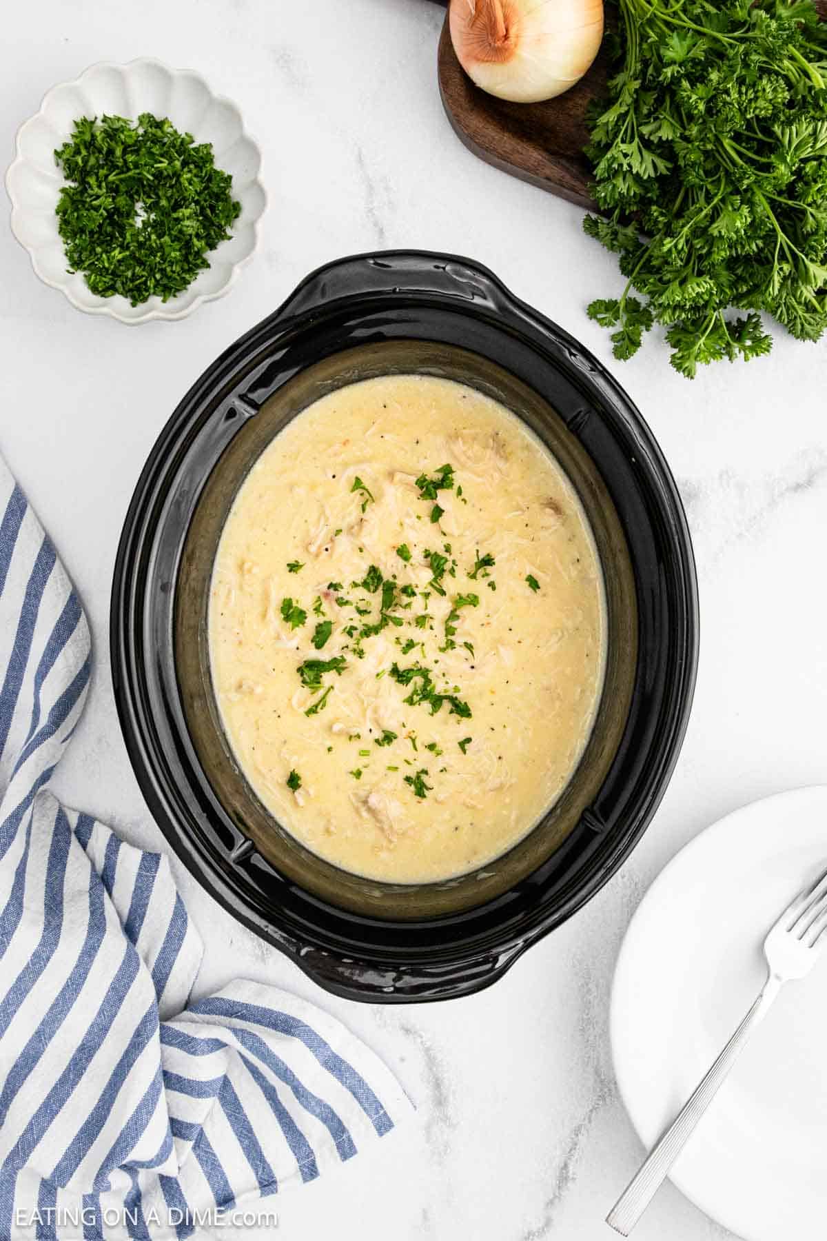 A creamy Crock Pot Chicken Alfredo and rice dish garnished with chopped parsley is shown in a black slow cooker on a marble countertop. Fresh herbs, a striped napkin, and a white plate with a fork are nearby.