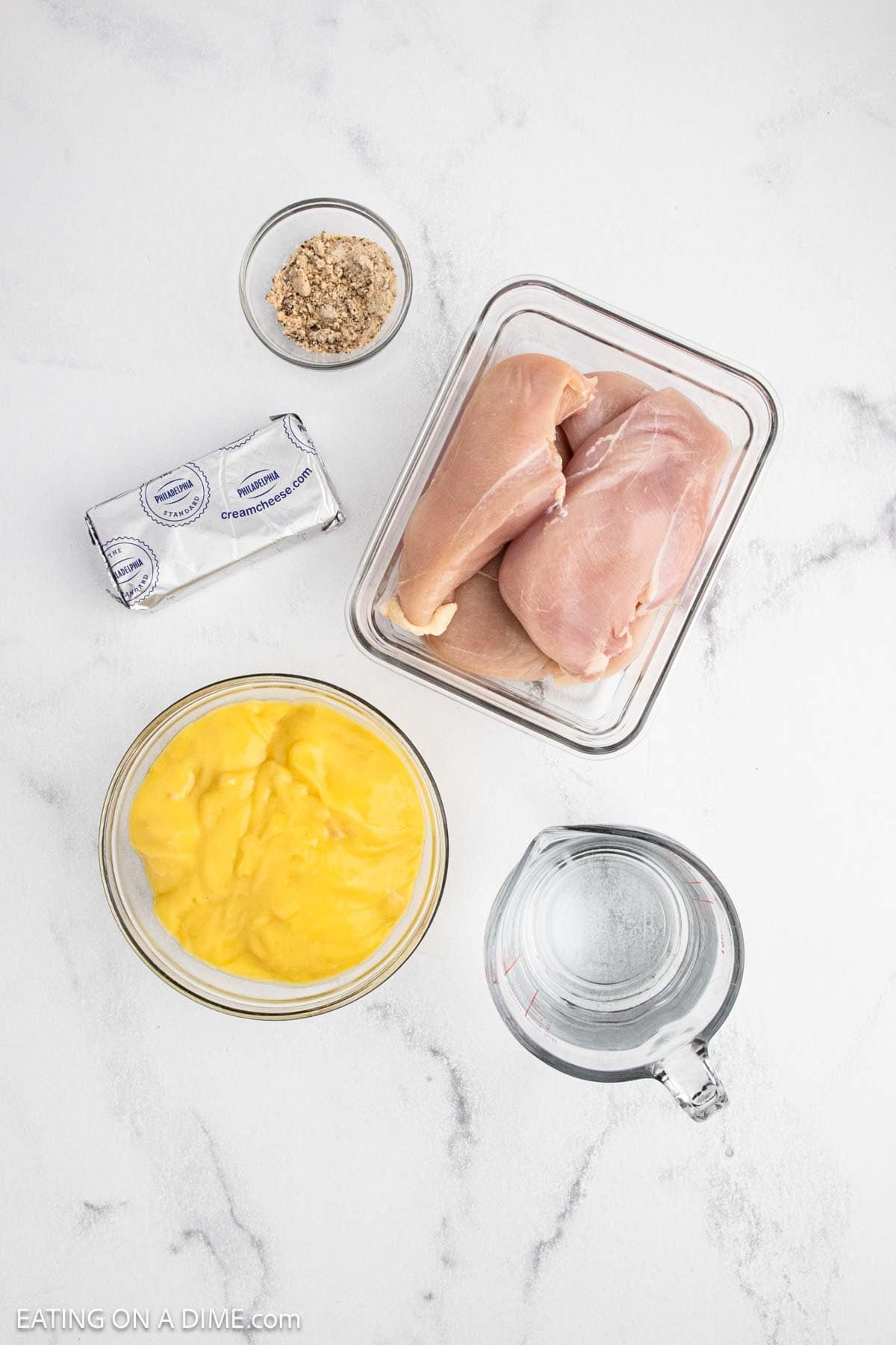 Overhead view of ingredients for Crock Pot Chicken Alfredo: raw chicken breasts in a glass container, a bowl of yellow soup, wrapped cream cheese, seasoning mix, and a measuring cup of water on white marble.