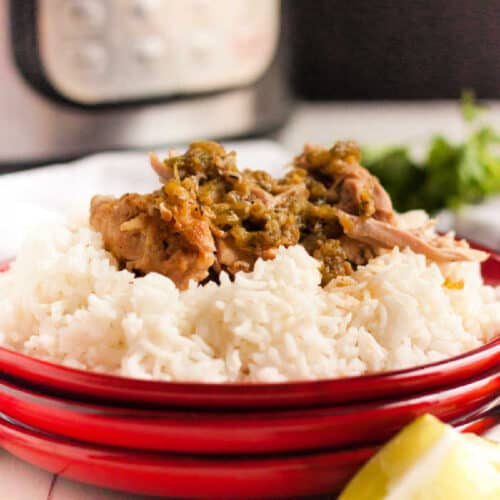A red plate holds a serving of white rice topped with shredded pork and green salsa, reminiscent of a zesty Instant Pot Salsa Verde Chicken recipe. In the background, there's a blurred pressure cooker and some cilantro. A lime wedge is positioned in the corner of the image.