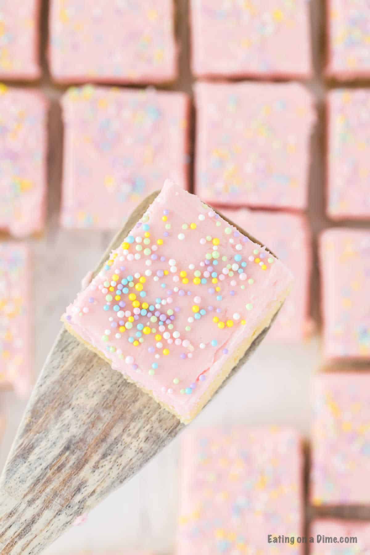 A wooden spatula holding a square piece of pink frosted cake with colorful sprinkles, reminiscent of delicious sugar cookie bars. Other similar cake squares are blurred in the background.