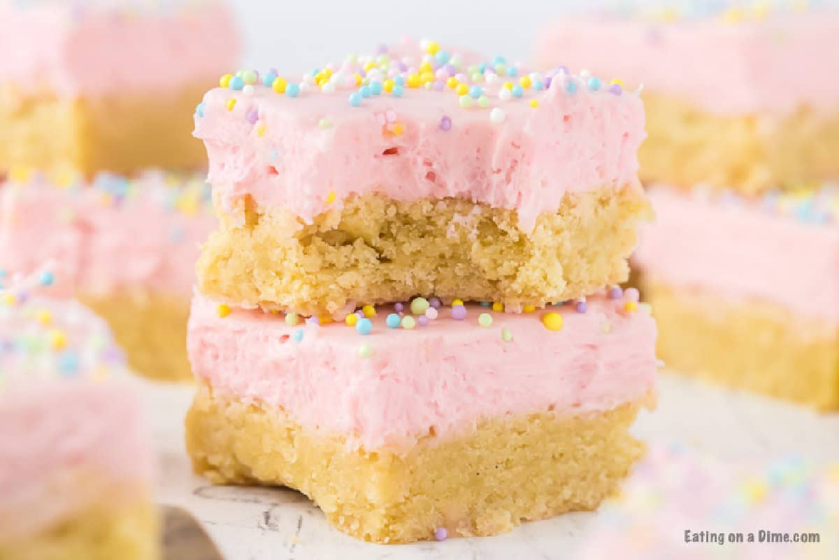 Two stacked Sugar Cookie Bars with thick pink frosting and colorful sprinkles on top. The top bar has a bite taken out, showing the soft texture inside. Other frosted Sugar Cookie Bars are blurred in the background.