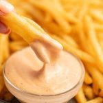 A hand dips a French fry into a creamy, orange-colored Freddy's Fry Sauce in a small glass bowl, with a pile of fries in the background.
