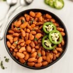 A black bowl filled with Instant Pot Pinto Beans, garnished with cilantro and topped with sliced fresh jalapeños, sits on a white surface with blurred utensils and jalapeño slices in the background.