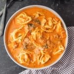A bowl of creamy Instant Pot Tortellini Soup with cheese tortellini, ground meat, and herbs sits on a checkered cloth with utensils nearby on a dark surface.