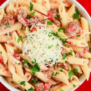 A bowl of penne pasta with roasted red pepper and Italian sausage, topped with shredded cheese and chopped parsley. The pasta is coated in a creamy sauce and served on a red background.