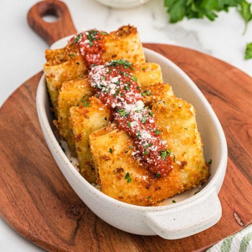 Four golden, crispy Olive Garden Fried Lasagna ravioli topped with marinara sauce and grated cheese, garnished with chopped parsley, served in a white oval dish on a wooden board.