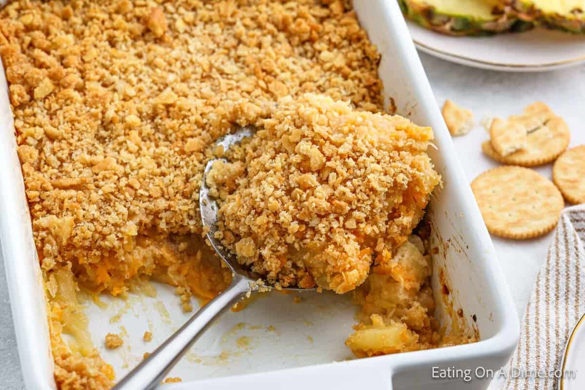 A white baking dish filled with a golden, crumbly pineapple casserole, with a serving spoon lifting a portion.