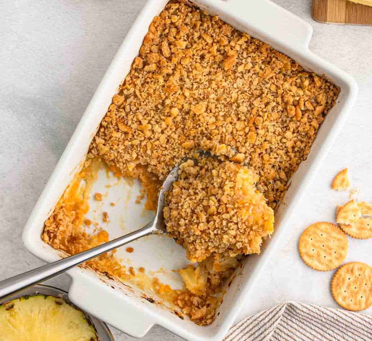 A rectangular baking dish contains a partially served pineapple casserole with a golden-brown, crumbly topping. A serving spoon rests inside the dish. Crackers partially crumble on the right, and a small piece of pineapple is visible in the bottom left corner.