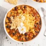 A bowl of 7 Can Taco Soup topped with crushed tortilla chips, sour cream, and shredded cheddar cheese. The soup features black beans, corn, tomatoes, and chicken. A striped napkin and spoon rest beside the bowl.