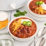 A bowl of turkey chili crockpot recipe topped with shredded cheddar cheese, sour cream, and sliced jalapeños sits on a white table beside two spoons, a napkin, and small bowls of extra cheese and sour cream. Two more bowls of chili are in the background.