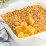 A white baking dish filled with Paula Deen Pineapple Casserole is topped with a golden crumbly crust. A serving spoon lifts out a portion, revealing sweet pineapple chunks beneath the topping. A striped cloth rests nearby.