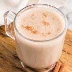 A clear glass mug filled with creamy, frothy horchata, reminiscent of a Starbucks Chai Tea Latte, is topped with a sprinkle of cinnamon and sits on a wooden surface with a cinnamon stick nearby.