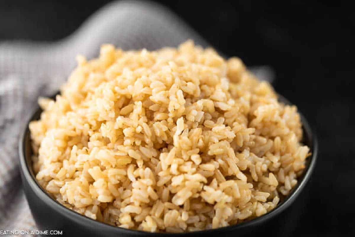 A black bowl brimming with fluffy, cooked brown rice sits elegantly on a dark surface. A soft beige cloth peeks into the background, adding a warm contrast to the scene—perfectly capturing a moment before discovering how to reheat rice flawlessly.
