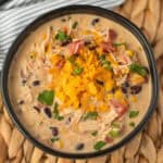 A bowl of Crockpot Creamy Chicken Taco Soup Recipe, topped with shredded cheddar cheese and chopped cilantro, is filled with black beans, corn, shredded chicken, and diced tomatoes. The bowl sits on a woven placemat.