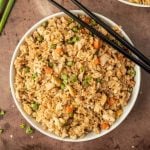 A bowl of chicken fried rice with peas, carrots, eggs, and green onions is presented in this easy Chicken Fried Rice Recipe, served alongside black chopsticks.