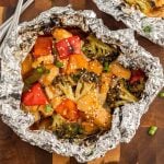 Foil packet filled with teriyaki chicken in foil, broccoli, red and yellow bell peppers, topped with sesame seeds and chopped green onions, placed on a wooden surface.