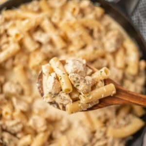 A wooden spoon holds a serving of spicy creamy chicken pasta, with tender chunks of chicken and fresh herbs, above a pan filled with more of this delicious pasta recipe.