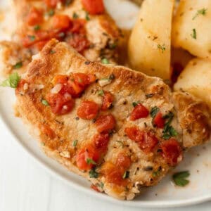 Pork chops on a white plate topped with diced tomatoes and chopped parsley.