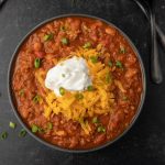 A bowl of Crock Pot Pumpkin Chili Recipe topped with shredded cheddar cheese, a dollop of sour cream, and chopped green onions, served on a dark surface with spoons in the background.