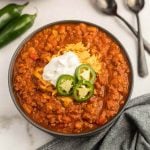 A bowl of pumpkin chili topped with shredded cheddar cheese, a dollop of sour cream, and sliced jalapeños, with two spoons and whole jalapeños in the background on a white surface.