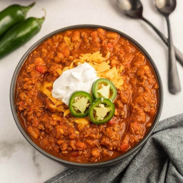 A bowl of pumpkin chili topped with shredded cheddar cheese, a dollop of sour cream, and sliced jalapeรฑos, with two spoons and whole jalapeรฑos in the background on a white surface.