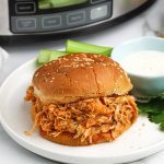 A Crock Pot Buffalo Chicken sandwich on a sesame seed bun is served on a white plate with celery sticks and a small cup of ranch dressing, with the slow cooker in the background.