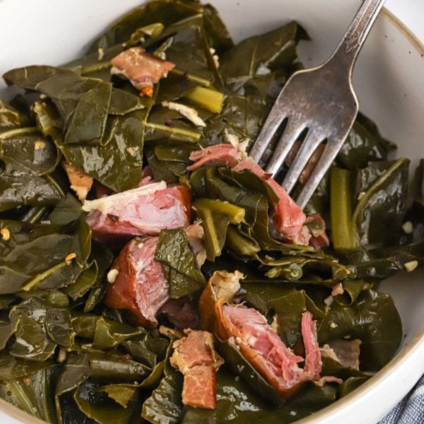 A bowl of Crock Pot Collard Greens mixed with pieces of smoked meat, with a fork resting on the side. The greens appear glossy and tender, and the meat is pink and shredded.